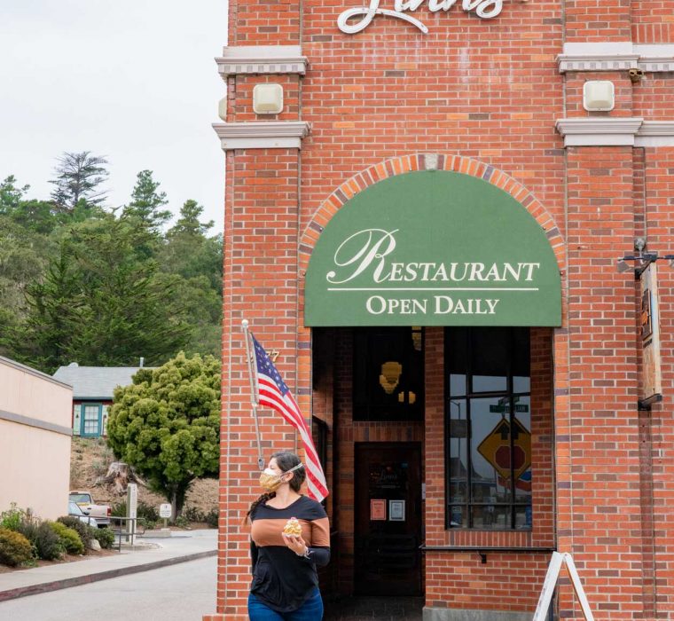A woman with a mask walking in a crosswalk across Main Street in Cambria, California, in front of Linn's Restaurant
