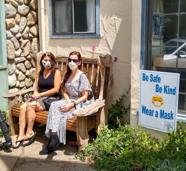 Two women sitting on a bench outside a shop, next to a sign with a masked face emoji saying "Be Safe, Be Kind, Wear a Mask" in Cambria, California