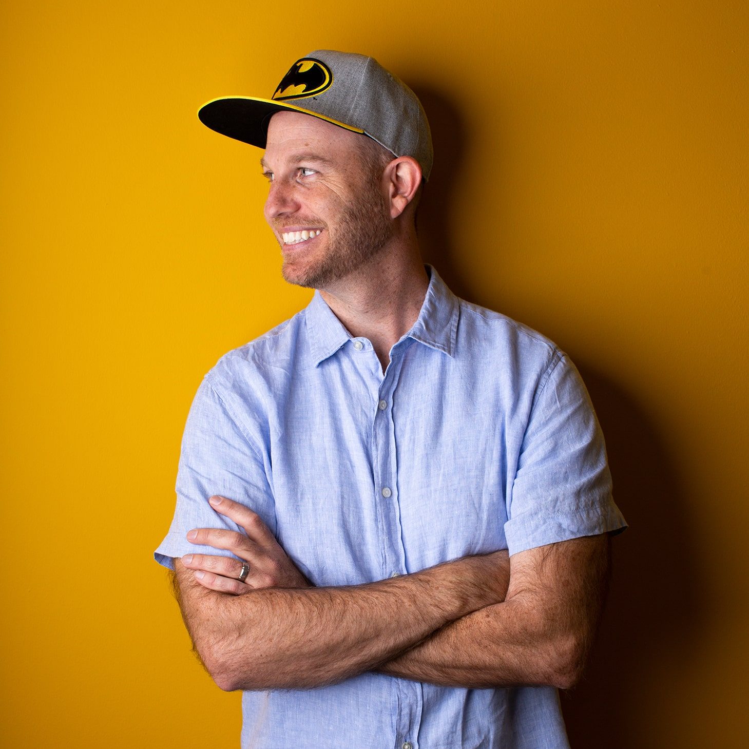 Profile of Gordon Howell in a Batman cap, smiling and looking to his right with hands folded on a dark yellow background