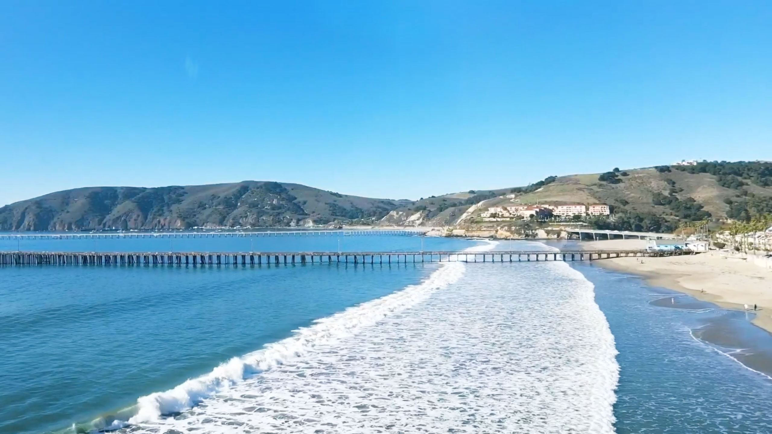 Pristine shot of a Central Coast beach, with a pier. extending from the beach to the left, past the photo into the ocean on the right, with green hills in the background and an open, blue sky